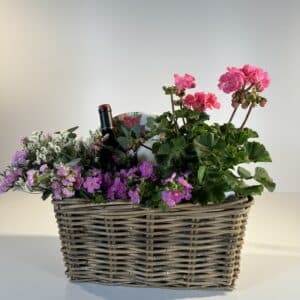 cesta de mimbre con gereanios, flores secas rosas y blancas, con una botella de vino blanco y una vela, ideal para hacer picnic.