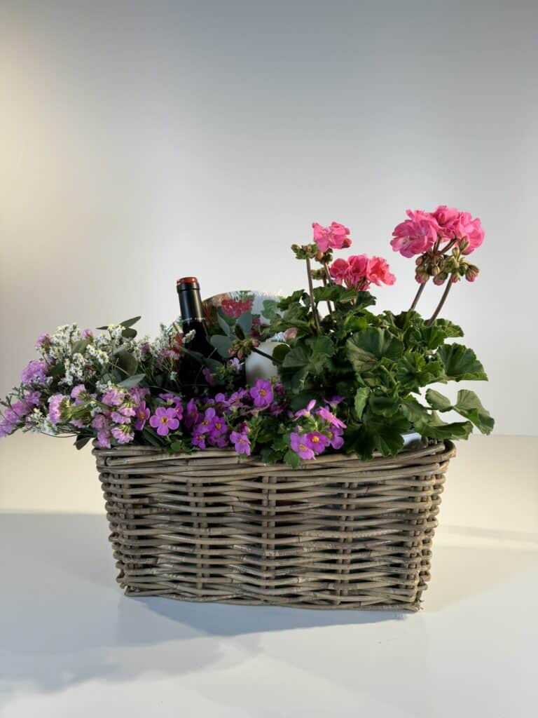 cesta de mimbre con gereanios, flores secas rosas y blancas, con una botella de vino blanco y una vela, ideal para hacer picnic.
