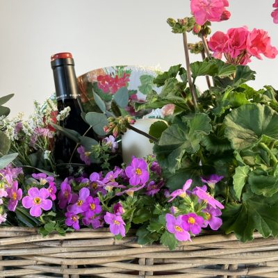 cesta de mimbre con gereanios, flores secas rosas y blancas, con una botella de vino blanco y una vela.