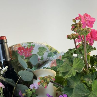 cesta de mimbre con gereanios, flores secas rosas y blancas, con una botella de vino blanco y una vela.