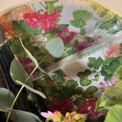 cesta de mimbre con gereanios, flores secas rosas y blancas, con una botella de vino blanco y una vela.