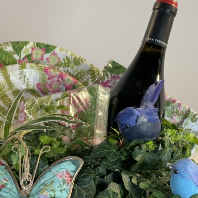 cesta de mimbre campestre con plantas verdes, platos servilletas, una botella de vino y adornos variados.